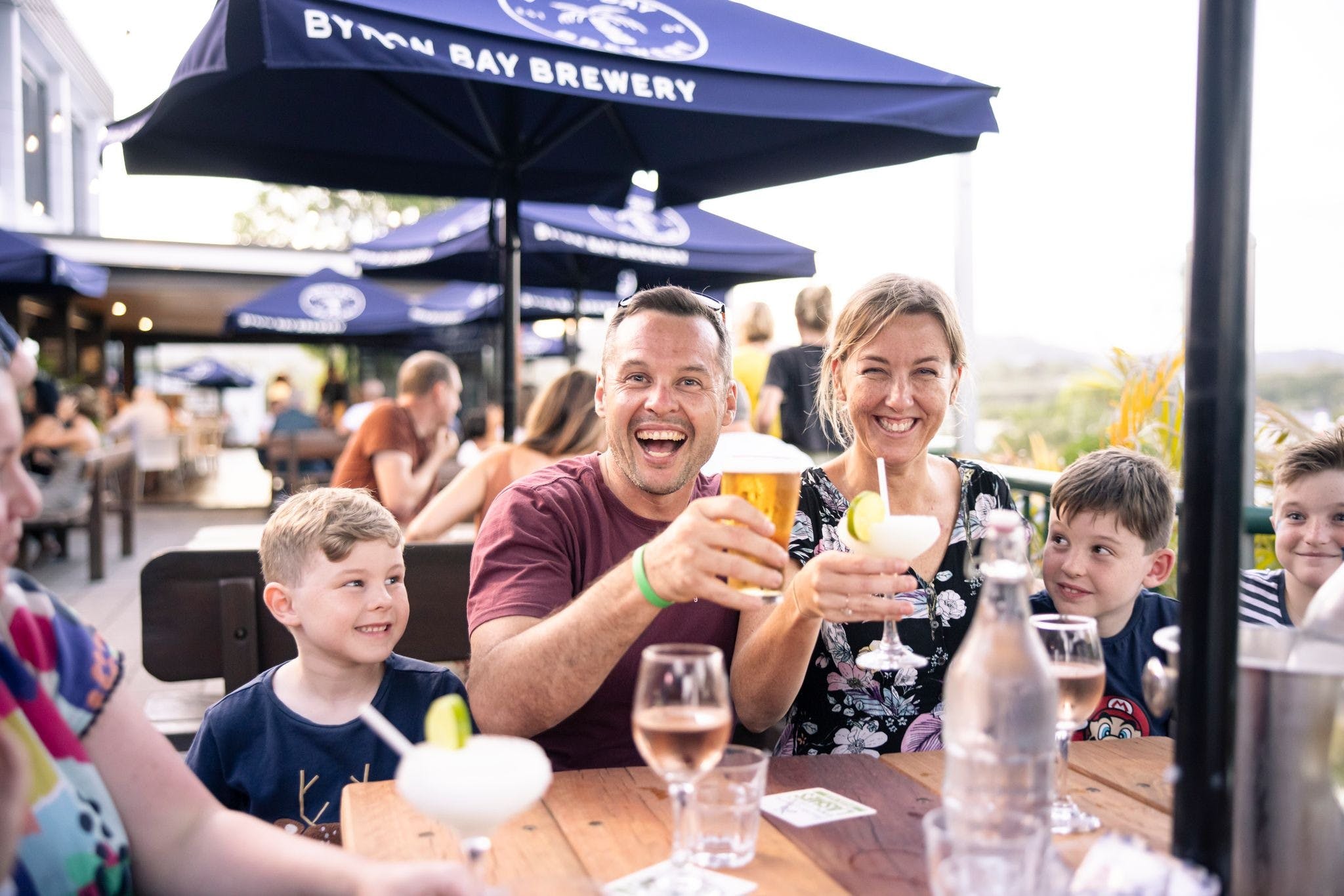 Mum, Dad and two kids sitting at an outdoor table smiling at the camera with drinks raised