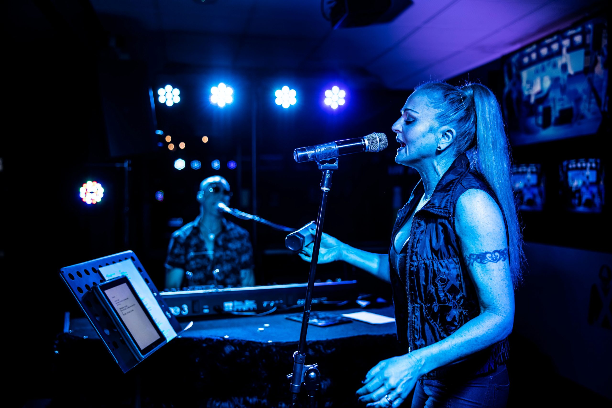 Woman in foreground singing into a mic with man in the background playing keyboard